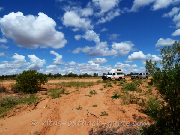 A campervan and another car on a dirt road in the Outback. Blue sky with fluffy white clouds, the sandy roads with green bushes, these are the colours of Australia